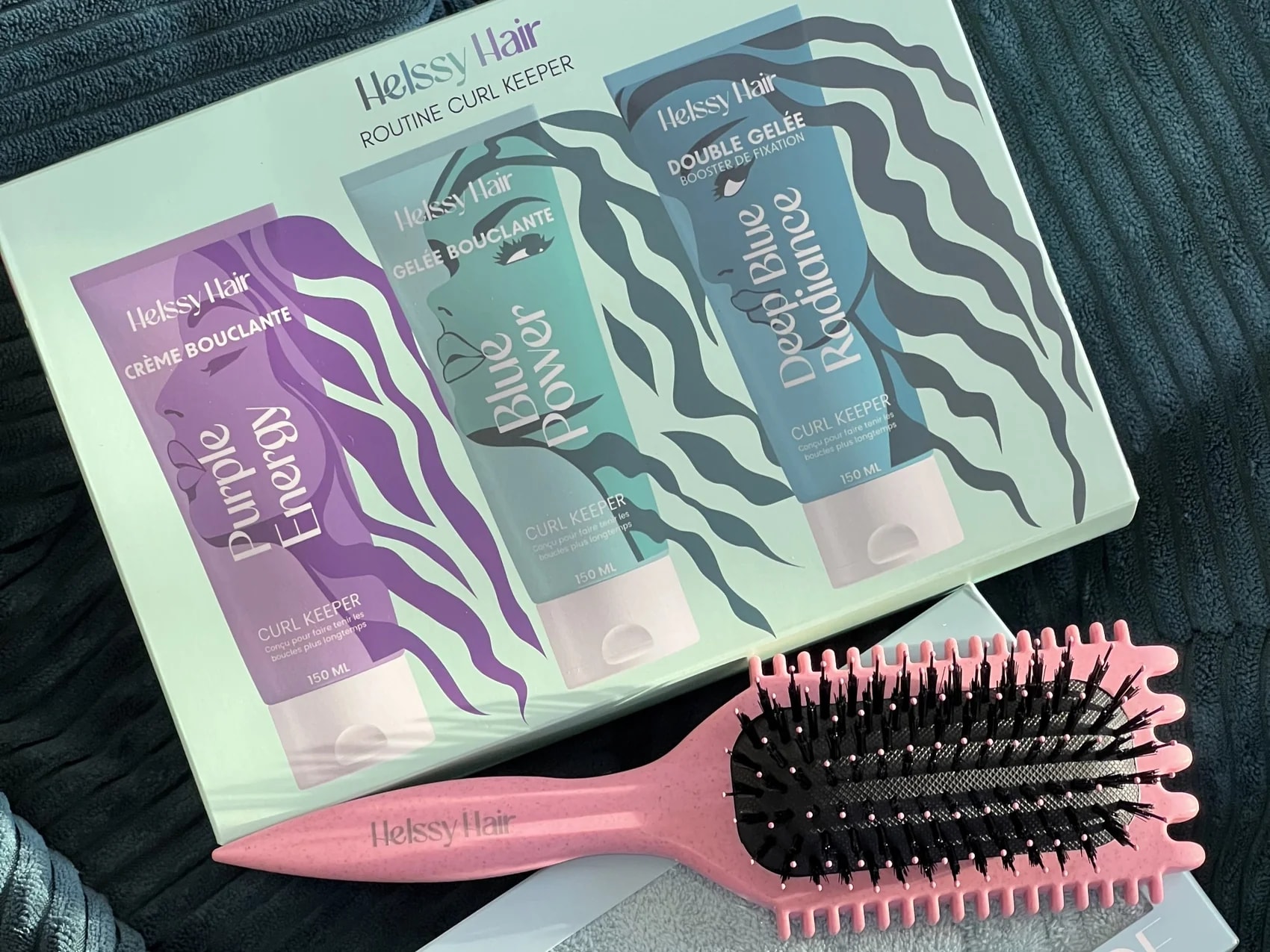 Le coffret Routine Curl Keeper de Helssy Hair, une brosse rose et une serviette microfibre posés sur un plaid côtelé bleu-vert.